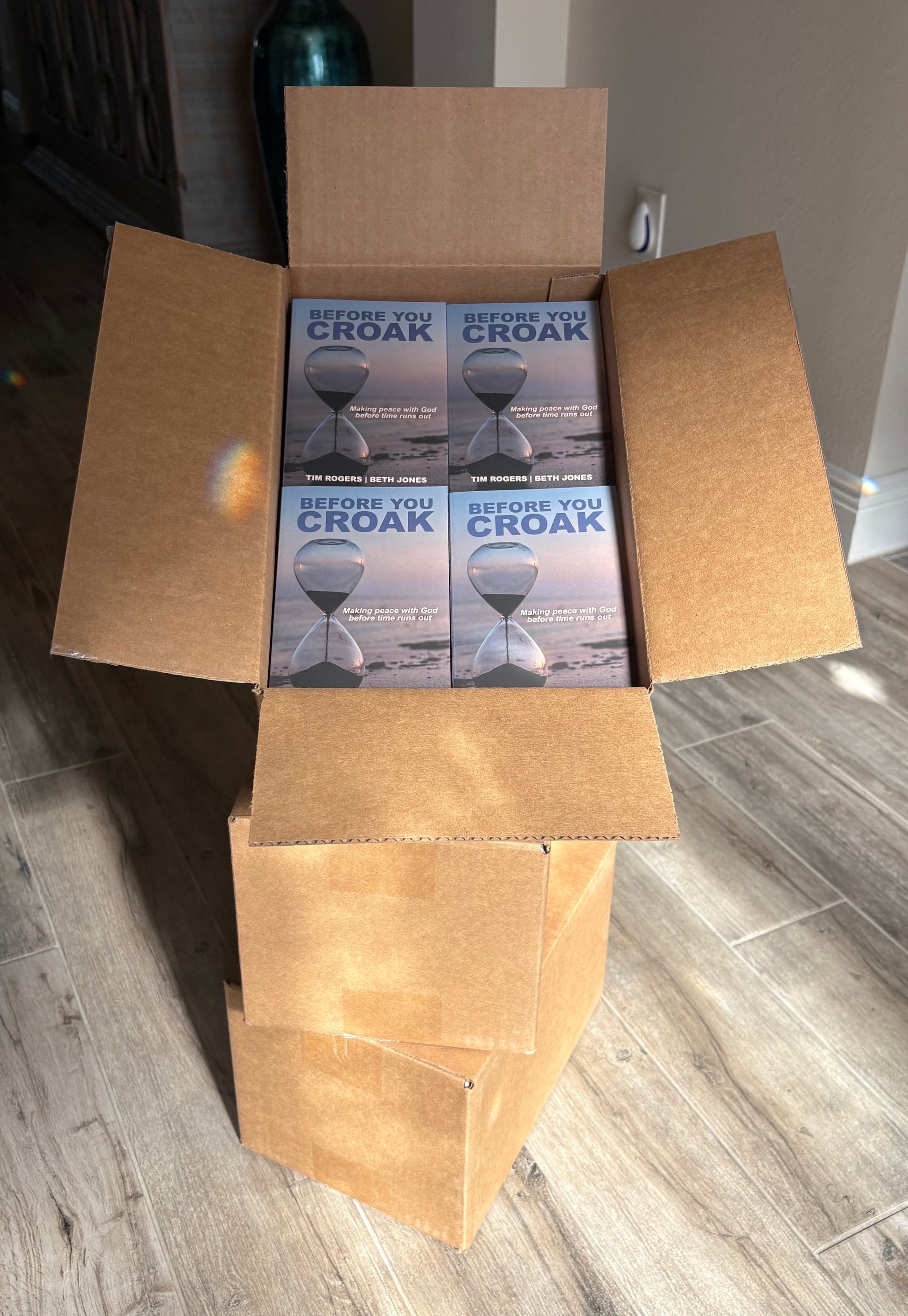 Open cardboard box containing multiple boxes of 'Before You Croak' products on a wooden floor.